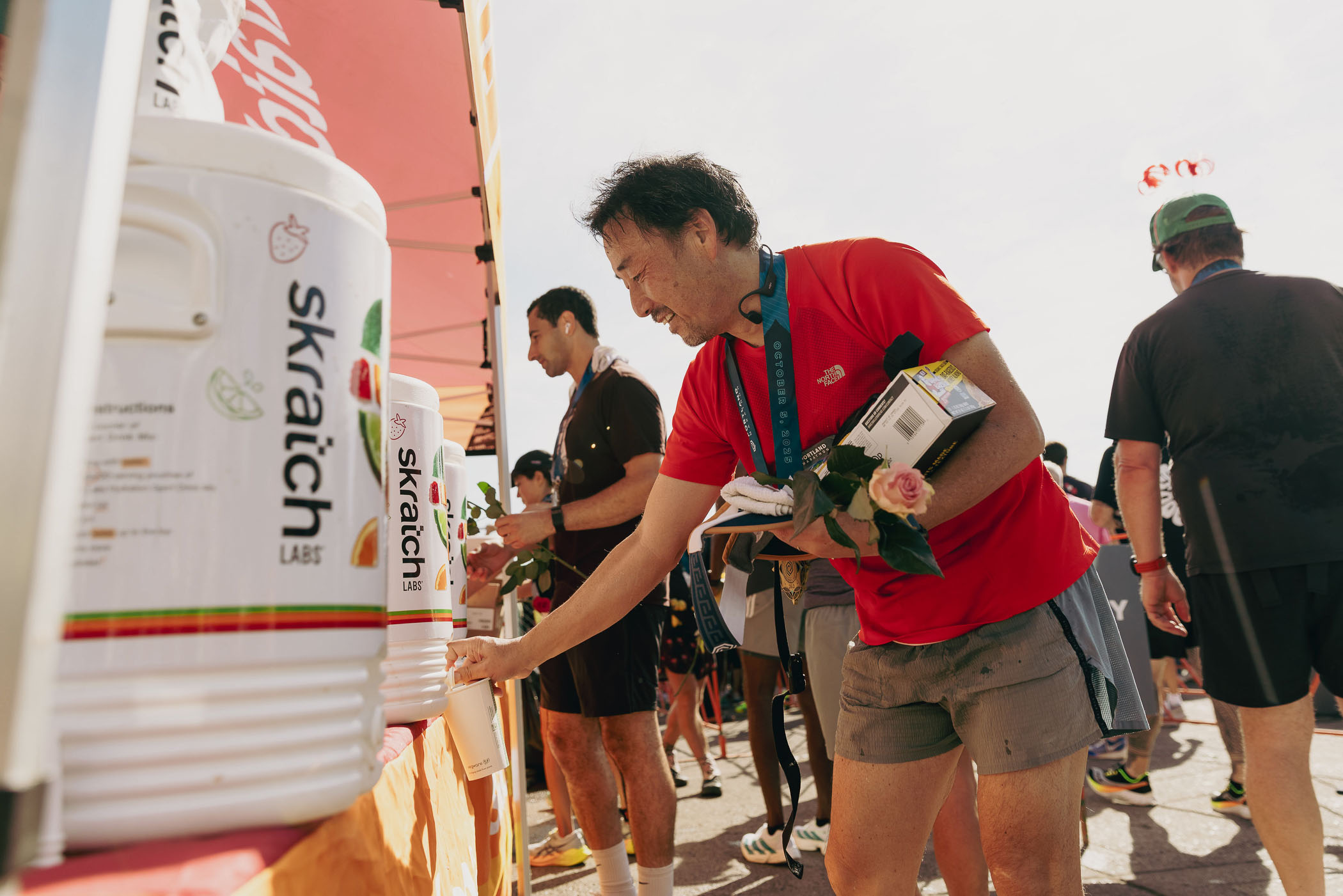 A man in a red shirt fills a cup at a Skratch Labs drink station during an outdoor event. He holds a rose, a box, and a medal, with other participants gathered under a tent nearby. Darby Communications PR + Digital Marketing